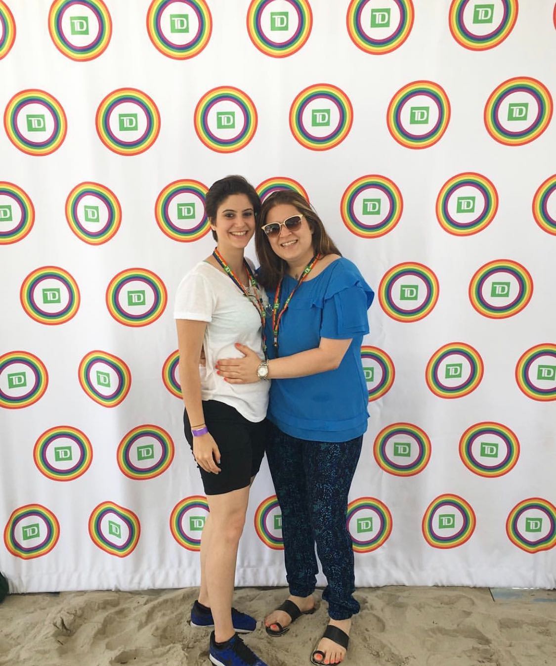 Christina Papadimitriou at the Long Beach PRIDE Parade with her girlfriend; Shows two women smiling, standing against photo backdrop