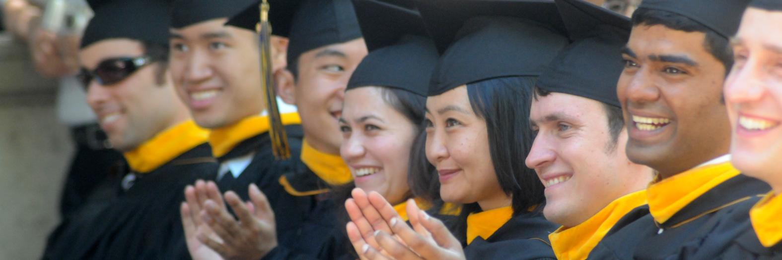 A group of graduating master's students at their commencement ceremony, wearing black caps and gowns with gold master's hoods.