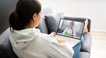 image of a person on a couch holding a laptop with someone wearing a physician's coat on the screen, and icons indicating a call