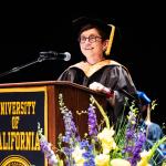 woman with glasses wearing graduation regalia speaking at a lectern
