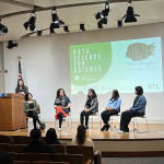 5 women seated on chairs on a stage and one woman at a podium. Behind them is a projected slide reading "data science for science" with the Women in Data Science Berkeley logo