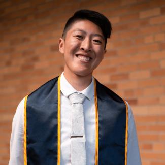 Kai standing against red brick wall while wearing a white button up and navy blue graduation stole. He is smiling with his head tilted slightly.