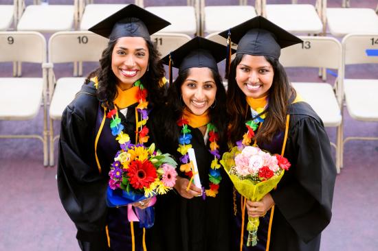 Three graduates wearing leis and graduation regalia pose together smiling and holding flowers