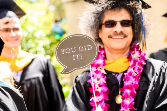a man in sunglasses, a lei, and graduation regalia holds a sign reading "You did it"