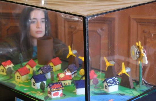 A student peers into a glass box with tiny model houses, windmills, and sun.
