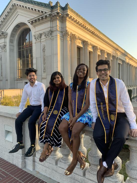four graduate students sit in front of the UC Berkeley library wearing graduation sashes
